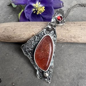 Kamień słoneczny granat wisior. Artystyczny srebrny wisior próby 925 z naturalnym kamieniem słonecznym w kształcie łzy, oprawionym w fakturowaną, asymetryczną trójkątną ramę z mocno oksydowanego i polerowanego srebra. Powyżej, na krawatce, osadzony jest mały, okrągły granat o średnicy 4 mm. Wisior marki Kamyki Moniki zaprezentowany na jasnym, gładkim tle. Obok biżuterii leży naturalny kawałek drewna, ukazujący przez kontrast surową fakturę srebra, oraz świeży kwiat, którego barwa podkreśla i eksponuje głęboki, winny kolor granatu oraz ciepłe refleksy kamienia słonecznego.