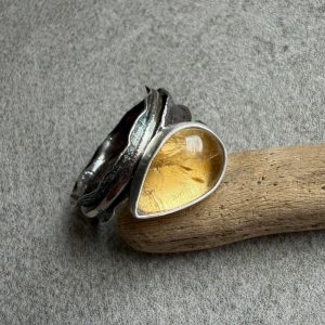 Cytryn pierścionek kropla. Artystyczny, szeroki pierścionek srebrny damski próby 925 z naturalnym cytrynem o ciepłej, żółtej barwie i kształcie smukłej kropli. Kamień ma wymiary 15 na 10 milimetrów i wysokość 6 milimetrów, osadzony w prostej, gładkiej oprawie. Konstrukcja opiera się na unikalnej, fakturowanej i falowanej obrączce, która pod kamieniem osiąga szerokość 10 milimetrów, a następnie asymetrycznie zwęża się ku końcowi, organicznie okalając palec niczym ogonek jaszczurki. Obrączka jest przecięta, co umożliwia swobodną regulację rozmiaru. Srebro ma surowy charakter, zostało głęboko zaoksydowane na ciemnoszary kolor, a następnie starannie przetarte, by uwypuklić rzeźbiarską strukturę metalu. Pierścień sfotografowany z bliska na jasnym tle jako autorski unikat handmade marki Kamyki Moniki.