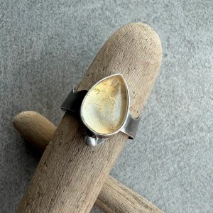 Cytryn pierścionek kropla . Artystyczny pierścionek srebrny damski próby 925 z naturalnym, przejrzystym cytrynem o ciepłej, miodowo-żółtej barwie. Kamień ma owalny kształt o wymiarach 17 na 12 milimetrów i wysokość 7 milimetrów, z czego spora część wystaje ponad gładką, srebrną oprawę. Pod minerałem znajduje się jasne srebro, które odbija światło i potęguje blask. Oprawa zamontowana jest prostopadle na szerokiej, 7-milimetrowej obrączce z funkcją regulacji rozmiaru, której jeden brzeg jest ozdobnie wycięty w kształt fali. Przy szerszej części kamienia widoczna jest mała, dekoracyjna kulka ze srebra. Srebro jest delikatnie oksydowane i wypolerowane na wysoki połysk. Pierścień sfotografowany z bliska na jasnym tle jako autorski projekt marki Kamyki Moniki.