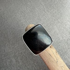 Pierścionek turmalin czarny duży. Artystyczny, monumentalny pierścionek srebrny damski próby 925 z dużym, naturalnym czarnym turmalinem schorl o geometrycznym kształcie zbliżonym do prostokąta. Kamień o imponujących wymiarach 27 na 25 milimetrów posiada naturalną fakturę z widocznymi, drobnymi rysami i mikrospękaniami strukturalnymi, charakterystycznymi dla tego minerału. Turmalin jest precyzyjnie zakuty w gładką oprawę ze srebra. Całość osadzona została na szerokiej, gładkiej obrączce o szerokości 6 milimetrów. Srebro poddano mocnemu oksydowaniu na ciemny kolor, a następnie zostało ono przetarte i wypolerowane, przez co ciemna patyna pozostała w zagłębieniach metalu, tworząc trójwymiarowy efekt retro. Obrączka ma konstrukcję przeciętą, co umożliwia swobodną regulację rozmiaru, również na większe palce. Pierścień sfotografowano z bliska na jasnym, minimalistycznym tle jako unikatowa biżuteria handmade marki Kamyki Moniki.