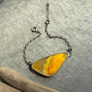 Artystyczna bransoletka srebrna próby 925 z dużym, naturalnym jaspisem trzmielim (bumblebee jasper) o wymiarach 38 na 19 milimetrów. Kamień ma unikalny kształt zaokrąglonego trójkąta i niesamowitą, warstwową barwę w paski o odcieniach intensywnej żółci, bieli oraz ciemnych linii. Minerał jest oprawiony w gładką, pełną ramkę ze srebra, która od spodu całkowicie zakrywa kamień. Srebrna oprawa jest lekko przyciemniana, oksydowana i wykończona na szlachetny, surowy mat. Bransoletka ma subtelny łańcuszek z regulacją długości i jest sfotografowana z bliska na jasnym tle jako unikatowa biżuteria handmade marki Kamyki Moniki.