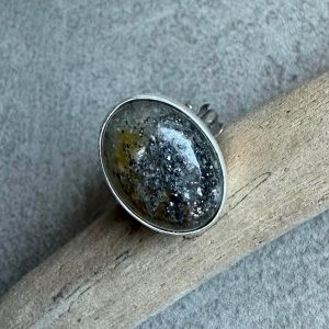 Pierścionek słoneczny kamień owal. Owalny pierścionek z czarnym kamieniem słonecznym ze srebra 925 na dłoni. Czarny kamień słoneczny owal 22x15 mm z efektem brokatowym. Falowana i regulowana srebrna obrączka pierścionka z czarnym kamieniem słonecznym. Stalowo-srebrna oprawa 925 wokół owalnego czarnego kamienia słonecznego